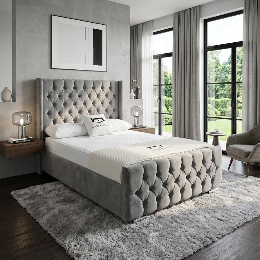 A modern bedroom featuring a gray tufted velvet bed frame with white bedding, set on a shaggy gray rug in a contemporary room with dark wood floors and large windows.