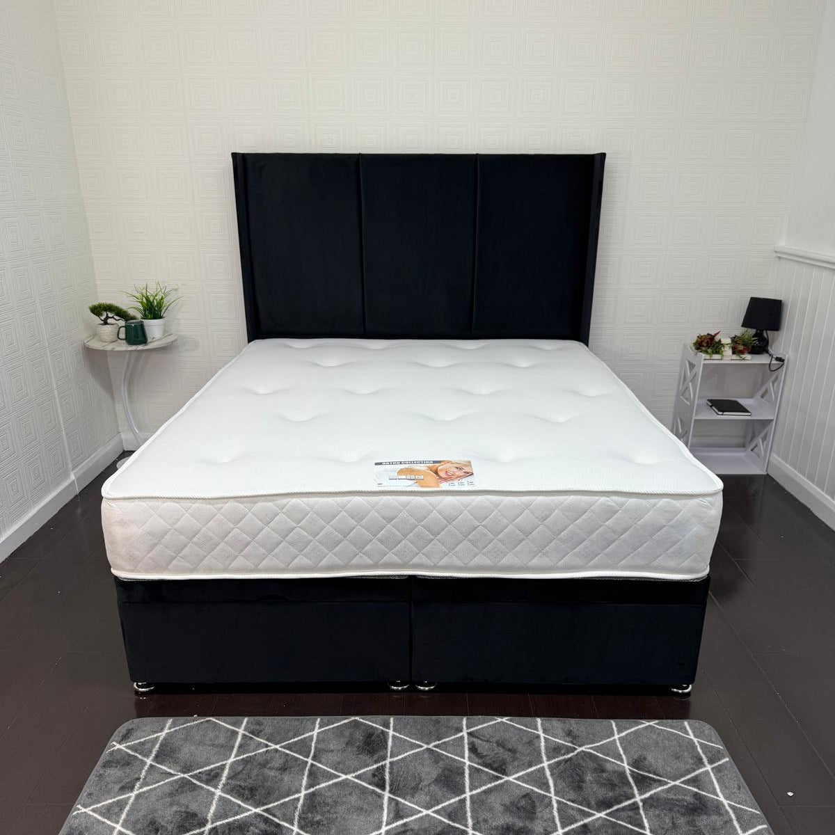 Bed with black headboard and white mattress in a room with a geometric patterned rug.