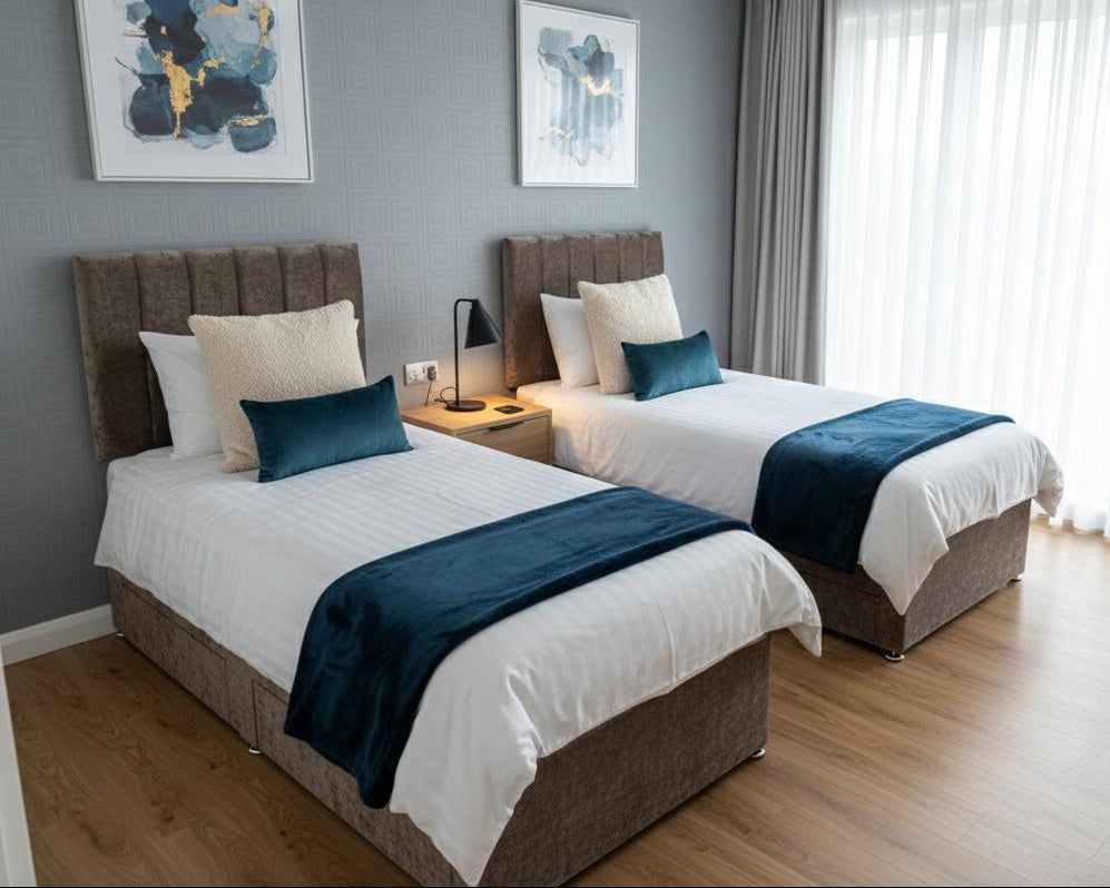 Two neatly made beds in a modern bedroom with blue accents and large windows.