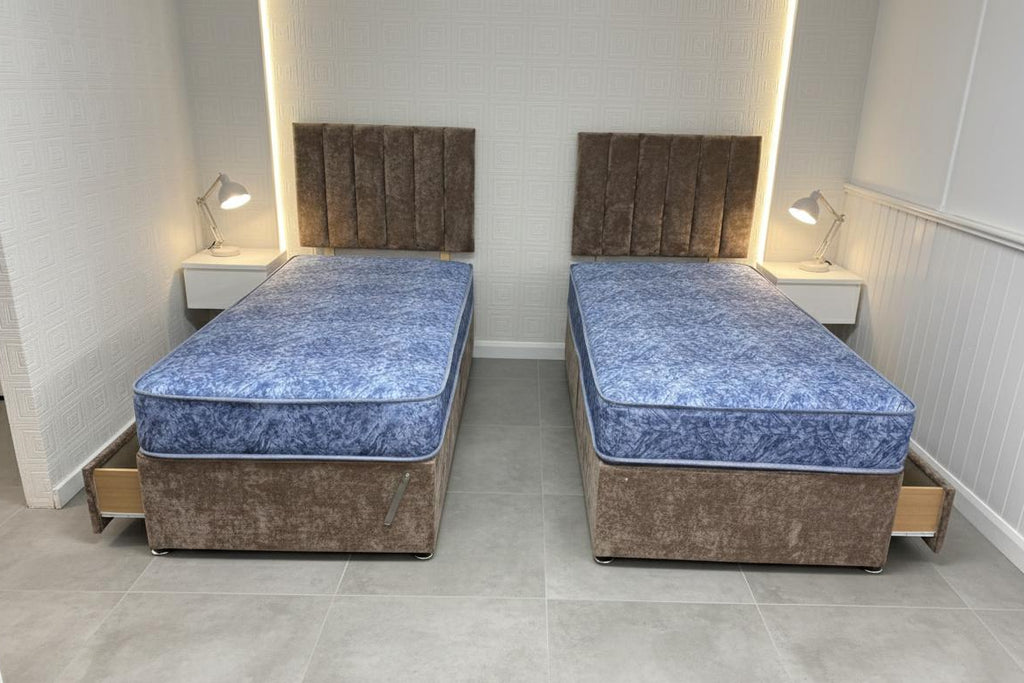 Two identical beds with blue mattresses in a room with white walls and gray flooring.