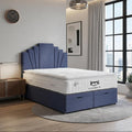 A modern bedroom scene showing a bed with a white mattress and a blue upholstered headboard and storage base, placed in a bright apartment interior with a large window, curtains, wall art, and a side table.