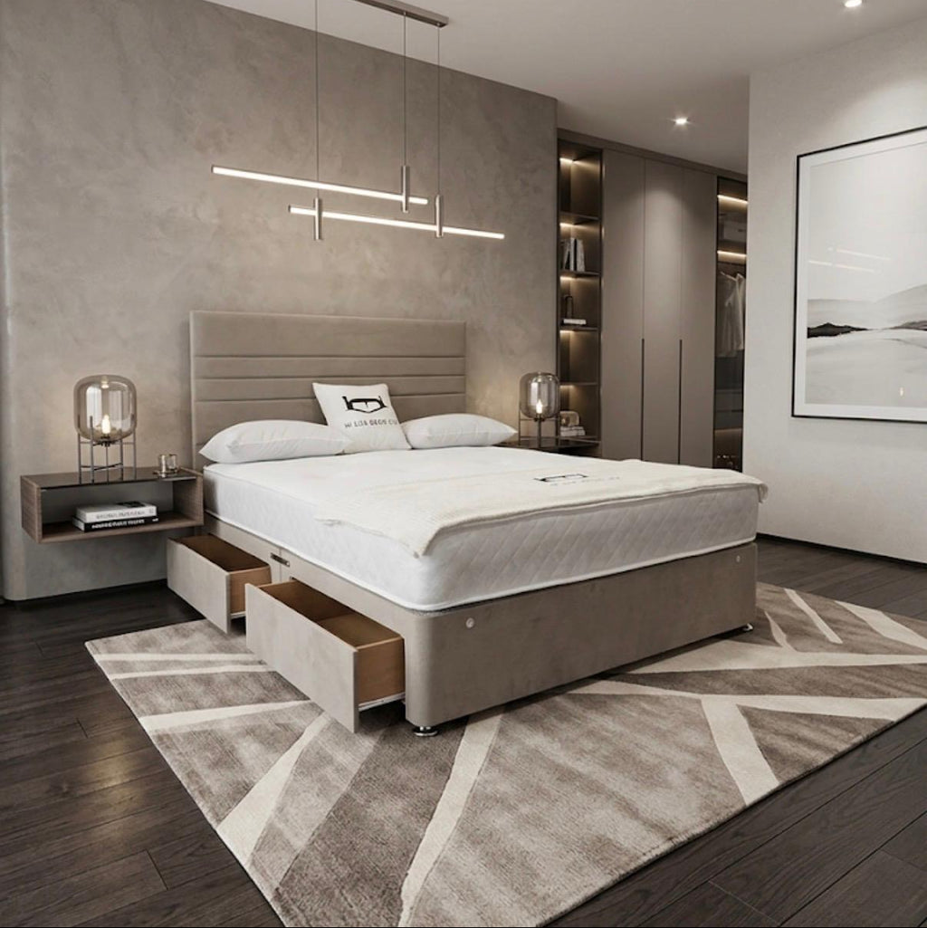 A modern bedroom featuring a low-profile platform bed with a light beige upholstered frame, white bedding, and an open storage drawer beneath the foot of the bed. The room has a textured gray accent wall, dark wood flooring, a geometric patterned rug, and contemporary linear pendant lighting.
