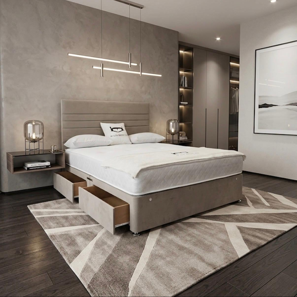 A modern bedroom featuring a low-profile platform bed with a light beige upholstered frame, white bedding, and an open storage drawer beneath the foot of the bed. The room has a textured gray accent wall, dark wood flooring, a geometric patterned rug, and contemporary linear pendant lighting.