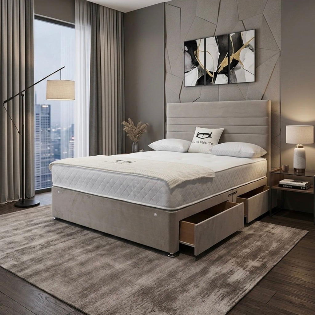 A modern bedroom featuring a beige upholstered bed frame with a quilted white mattress, matching headboard, and storage drawers. The room has a textured gray accent wall with a geometric abstract painting, a large window with city views, and a gray area rug.