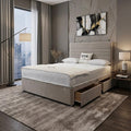 A modern bedroom featuring a beige upholstered bed frame with a quilted white mattress, matching headboard, and storage drawers. The room has a textured gray accent wall with a geometric abstract painting, a large window with city views, and a gray area rug.