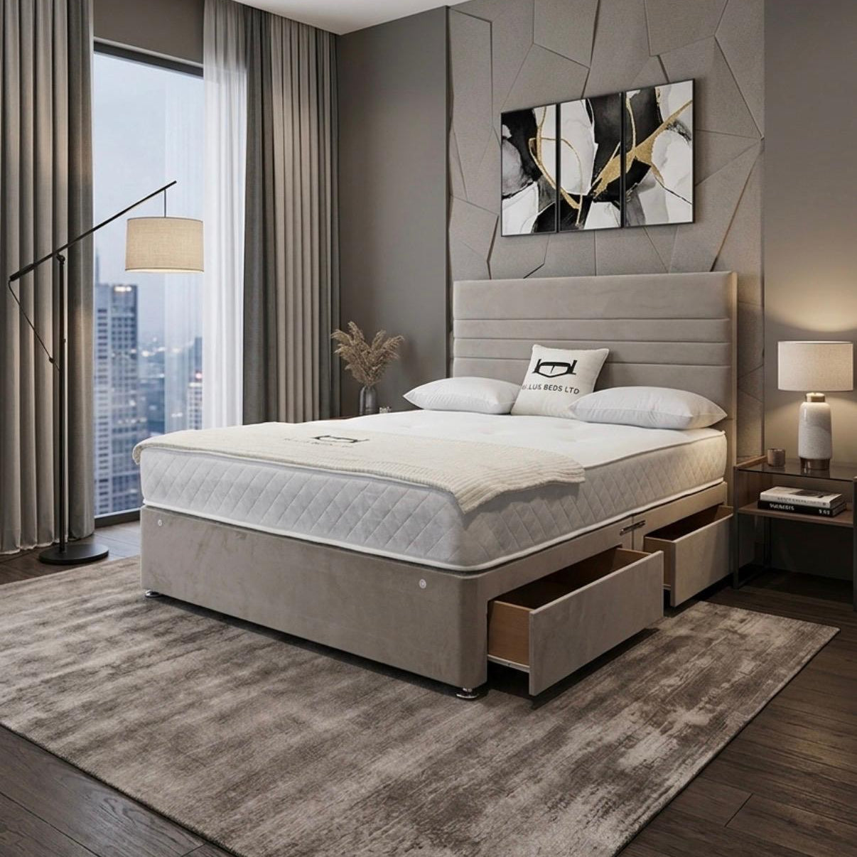 A modern bedroom featuring a beige upholstered bed frame with a quilted white mattress, matching headboard, and storage drawers. The room has a textured gray accent wall with a geometric abstract painting, a large window with city views, and a gray area rug.