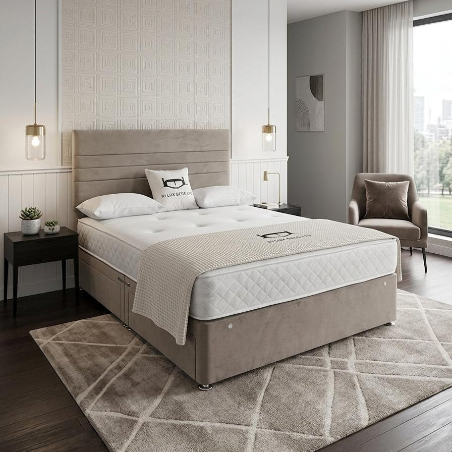A modern bedroom featuring a bed with a beige upholstered headboard and base, dressed in white and beige bedding. The bed is placed on a large geometric patterned rug over dark wood flooring, with a matching armchair and pendant lights nearby.