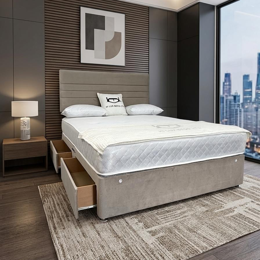 A modern bed with a light beige upholstered base and headboard, dressed in white bedding, set in a contemporary bedroom with dark wood floors, a patterned rug, and a large window overlooking a city skyline.