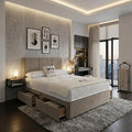 A modern bedroom featuring a low-profile platform bed with built-in storage drawers at the foot, dressed in white bedding. The bed is positioned on a large gray shag rug over dark wood flooring, with a textured beige accent wall, three framed black-and-white art prints, and a warm LED ceiling light above.