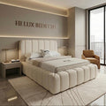 A luxury upholstered bed in a modern beige bedroom, featuring a segmented tufted headboard and matching footboard. The room includes a matching beige armchair, bedside tables with lamps, and a large window with a view of a city skyline.