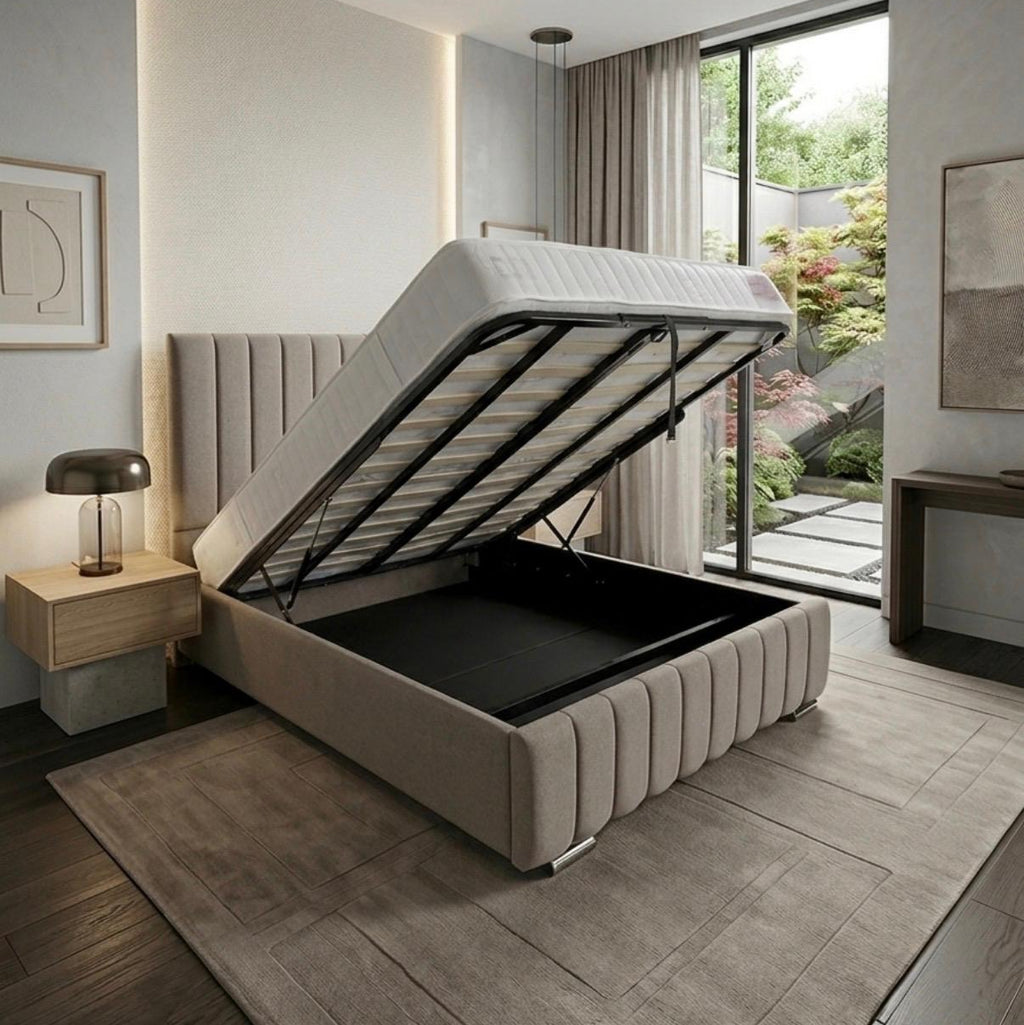 A modern upholstered storage bed with the mattress lifted open to reveal a black storage compartment, shown in a styled bedroom interior with a window, rug, and bedside lamp.