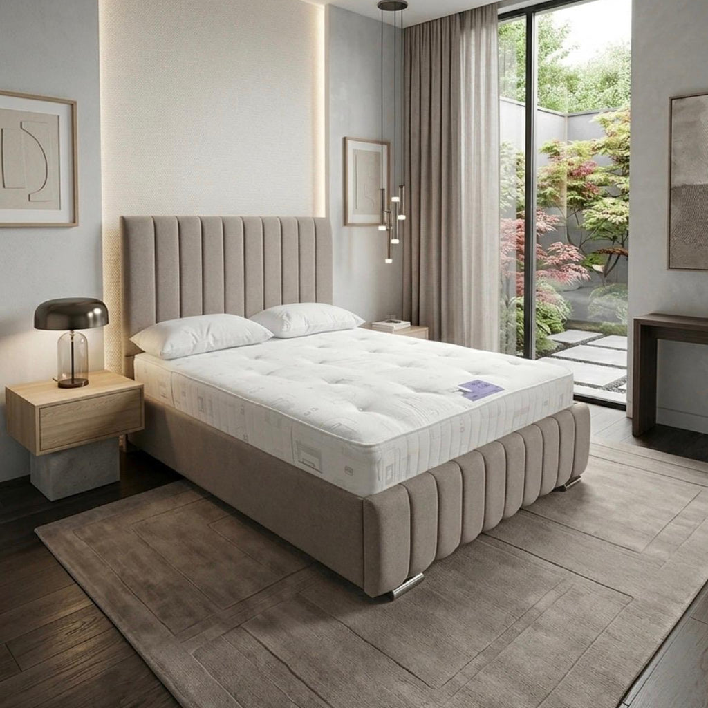 A modern upholstered bed with a tall channel-tufted headboard and matching footboard, shown in a styled bedroom setting with a white mattress, bedside tables, and a large window looking out to a garden.