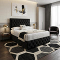 A modern bedroom featuring a black tufted upholstered bed frame with a white quilted mattress, styled with a black and white geometric area rug, bedside tables with gold lamps, and a dark gray armchair.