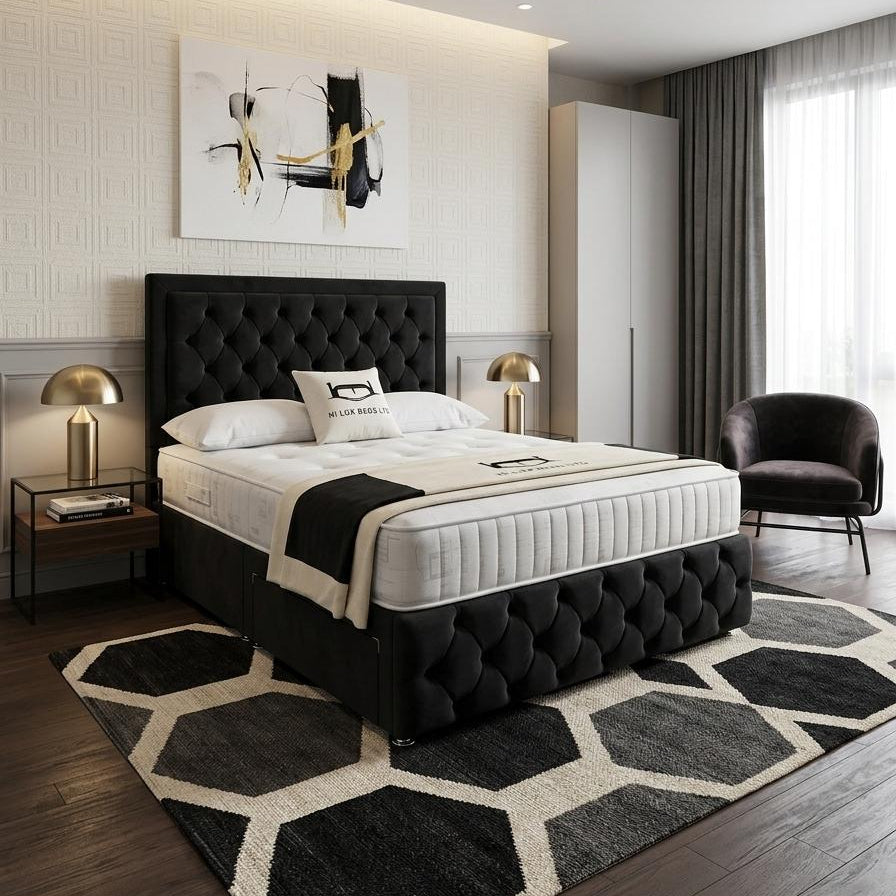 A modern bedroom featuring a black tufted upholstered bed frame with a white quilted mattress, styled with a black and white geometric area rug, bedside tables with gold lamps, and a dark gray armchair.