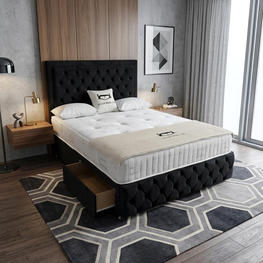 A modern bedroom setup featuring a tufted black bed frame with an open storage drawer underneath, dressed with a white tufted mattress and a beige quilted cover. The bed is placed on a dark gray and beige geometric patterned rug in a contemporary bedroom with wood flooring, a wood-paneled accent wall, and a large window.