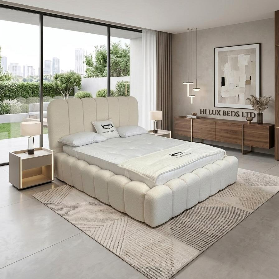 A modern upholstered platform bed in a bright, contemporary bedroom. The bed features a high, tufted headboard and matching tufted base in a light cream/off-white fabric, dressed with white bedding.