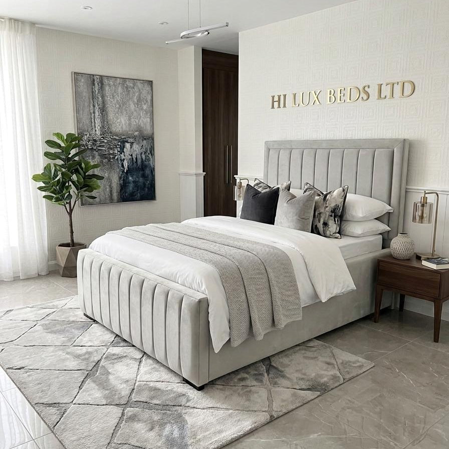 A modern bedroom featuring a light gray upholstered platform bed with a ribbed headboard and footboard, dressed in white and gray bedding. The room has a gray geometric pattern rug, a potted plant, and a large abstract painting on the wall.
