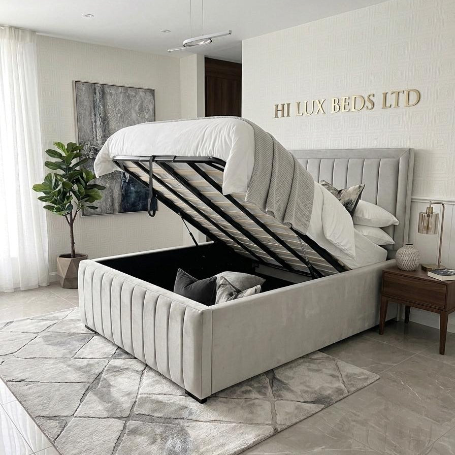 A light gray upholstered storage bed with its lift-up mattress platform open, shown in a modern bedroom setting. The room features a gray patterned rug, a wooden nightstand, and a plant, with the brand name 'HI LUX BEDS LTD' displayed on the wall behind the bed.