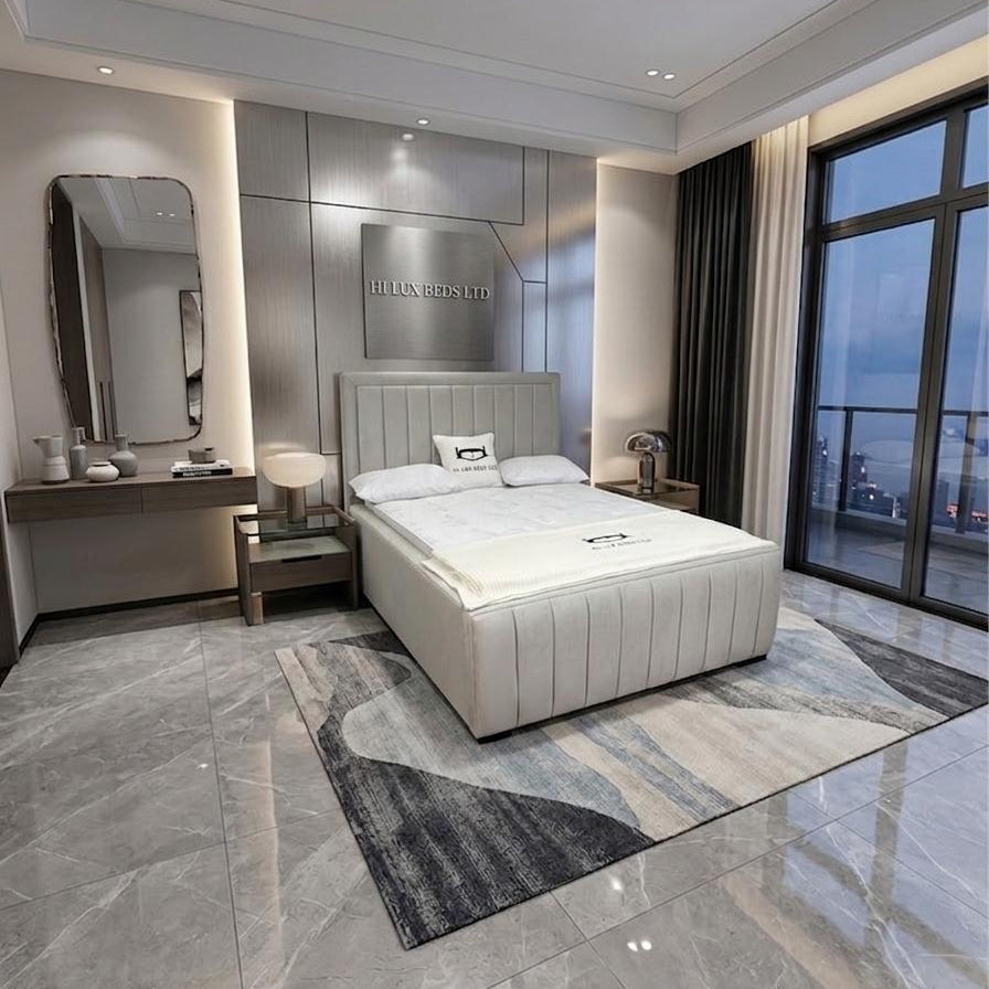 A modern bedroom featuring a cream tufted platform bed with white bedding, set on a gray and white abstract patterned area rug. The room has gray marble-look tiled floors, a matching gray vanity desk with a mirror, and a large floor-to-ceiling window looking out onto a city skyline at dusk.