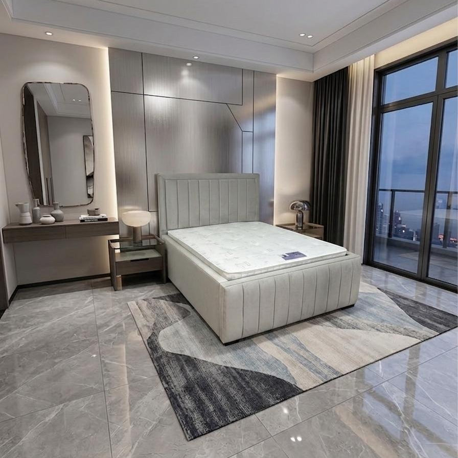 A modern bedroom featuring a light beige upholstered bed with a white mattress, set on a gray and beige patterned area rug. The room has gray marble-look tiled floors, a built-in vanity with a mirror, and large floor-to-ceiling windows with dark curtains.