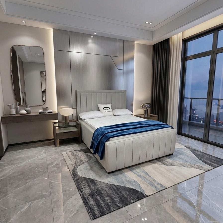 A modern bedroom featuring a light beige upholstered bed with white bedding and a blue throw blanket, set on a gray and white abstract patterned rug. The room has gray marble-look tiled floors, a built-in vanity with a mirror, and large floor-to-ceiling windows with a view of a city skyline at dusk.