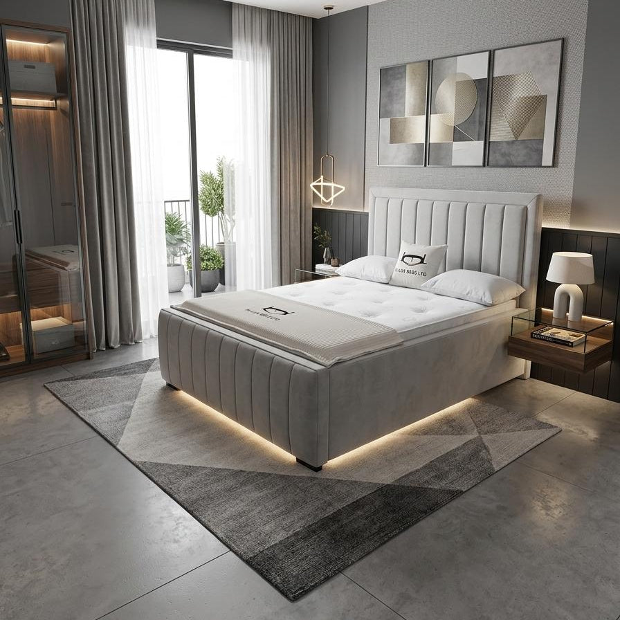 A modern bedroom featuring a light gray upholstered bed with a padded headboard and matching footboard, styled with white bedding and warm under-bed lighting. The room has gray walls, a geometric patterned rug, a large window with sheer curtains, and contemporary wall art.