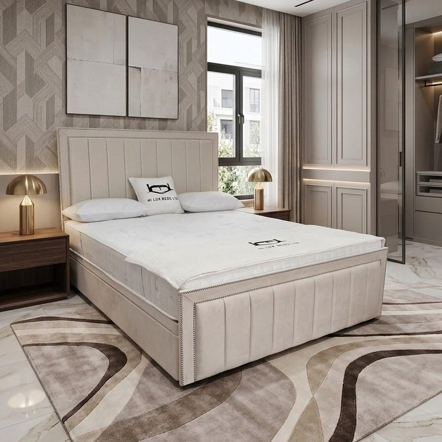 A modern bedroom featuring a beige upholstered bed frame with a white mattress, styled with neutral-toned bedding, a matching patterned area rug, and contemporary wall decor.